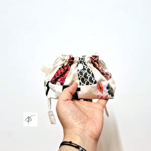 May include: A small, patchwork drawstring bag with a cream base and panels of patterned fabric in red, black, and white. The bag is held in a hand against a white background. The bag's drawstring is visible.