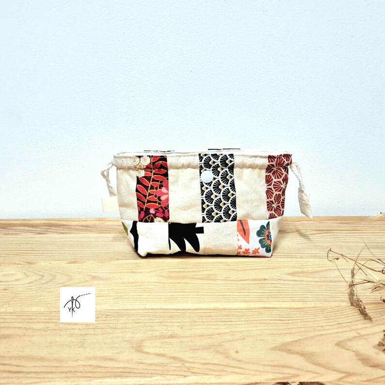 May include: A small, rectangular fabric basket with a patchwork design. The basket features panels of cream-coloured fabric with floral and geometric patterns in red, black, and green. It has small rope handles and is set on a wooden surface.