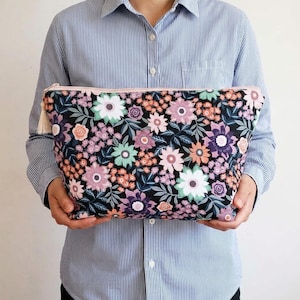 Pode incluir: Uma bolsa retangular com zíper em fundo preto com um denso padrão floral em tons de branco, rosa claro, lavanda e laranja suave, acentuado por folhas verde-escuras e um zíper rosa claro com um puxador de borla correspondente.