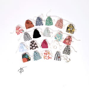 Könnte beinhalten: Eine Auswahl kleiner Stoffbeutel mit Kordelzug in verschiedenen Farben und Mustern, darunter Blumen-, geometrische und gestreifte Designs. Die Beutel sind auf einer weißen Oberfläche angeordnet, mit etwas Schmuck.