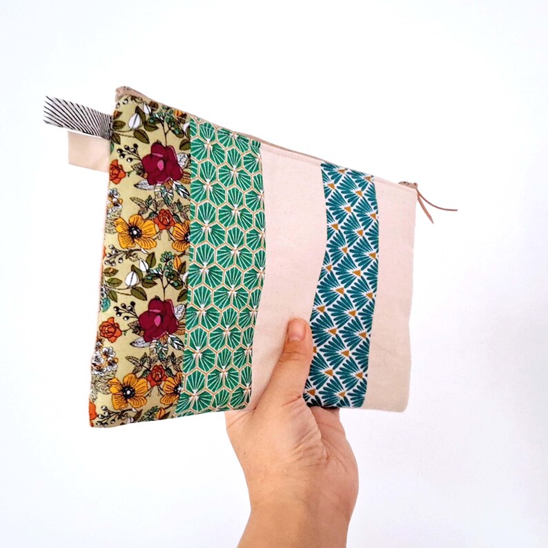 Op de afbeelding: Een rechthoekig stoffen etui met ritssluiting. De etui heeft een patchwork-ontwerp met bloemen-, geometrische en effen stoffen panelen in tinten groen, geel, roze en blauwgroen. Een gestreept lint en een leren trekker voegen details toe.