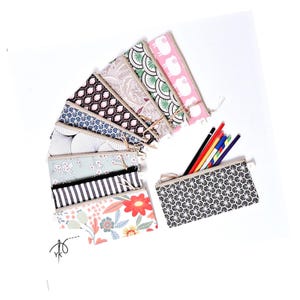 Könnte beinhalten: Eine Auswahl an Stoff-Federmäppchen mit verschiedenen Mustern, darunter Blumen, geometrische Formen und Schaf-Designs. Ein Etui ist geöffnet und zeigt Buntstifte. Die Etuis haben einen Reißverschluss und einen neutralfarbenen Besatz.