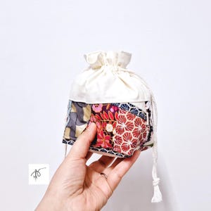 May include: A small drawstring pouch with a patchwork design. The lower portion features a mix of floral and geometric patterns in red, pink, blue, and gray. The top is a solid off-white fabric, and a white drawstring is visible.
