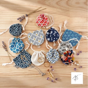 Könnte beinhalten: Eine Sammlung kleiner Stoffbeutel mit Kordelzug, die verschiedene geometrische, florale und fächerförmige Muster in Farben wie Rot, Blau, Grau und Creme aufweisen. Einige Beutel enthalten Goldringe, eine Silberkette und Goldflocken in einem Glasfläschchen.