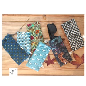 Peut inclure: Plusieurs étuis à lunettes en tissu aux motifs variés, dont des motifs floraux, de grues et géométriques. Un étui contient des lunettes de soleil noires. Les étuis sont bleus, verts et noirs, disposés sur une surface en bois.