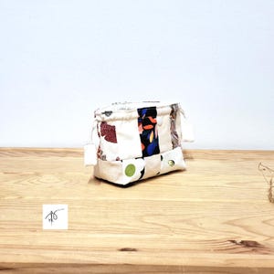 May include: A small, patchwork fabric pouch with a drawstring closure. The pouch is made of various fabric scraps in white, cream, and patterned fabrics with red, blue, and green accents. The pouch is sitting on a wooden surface.