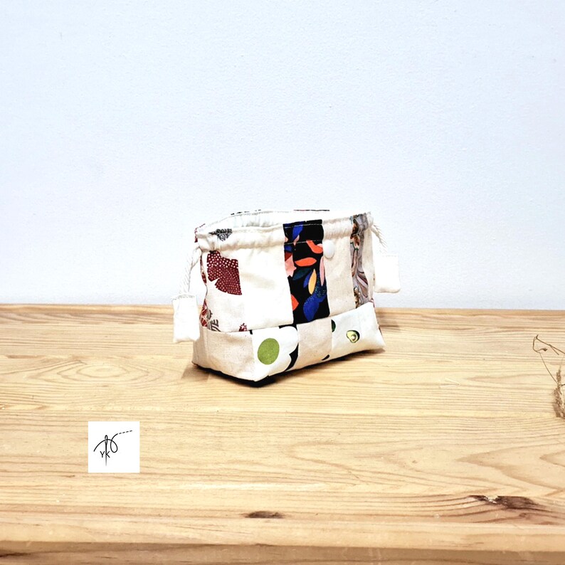 May include: A small, patchwork fabric pouch with a drawstring closure. The pouch is made of various fabric scraps in white, cream, and patterned fabrics with red, blue, and green accents. The pouch is sitting on a wooden surface.
