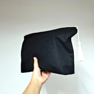 以下が含まれることがあります： 白いタッセルが付いた黒いクラッチバッグ。白い背景に、バッグは手で持たれています。クラッチは滑らかで不透明な素材で作られています。