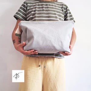 May include: A large, rectangular, light grey fabric pouch featuring a zip closure with a small, off-white decorative tassel attached to the side. The pouch has a smooth, solid-coloured surface and is held horizontally in front of the torso.