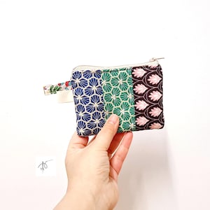 Può includere: Un piccolo portamonete rettangolare con chiusura a cerniera. Il portamonete è diviso in tre sezioni con diversi motivi geometrici blu, verdi e marrone scuro. Un piccolo anello di tessuto con un motivo floreale è attaccato al lato.