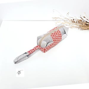 May include: A rectangular pencil case with a wrist strap. The case has a red and white geometric pattern on one side and a black and white fan-like design on the other. A beige zip runs along the top.