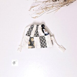 Op de afbeelding: Een kleine patchwork-zak met trekkoord. De tas is gemaakt van vierkante stoffen met patronen in zwart, goud en wit, tegen een witte achtergrond. Hij heeft een wit trekkoord en is vastgebonden met een wit koord.