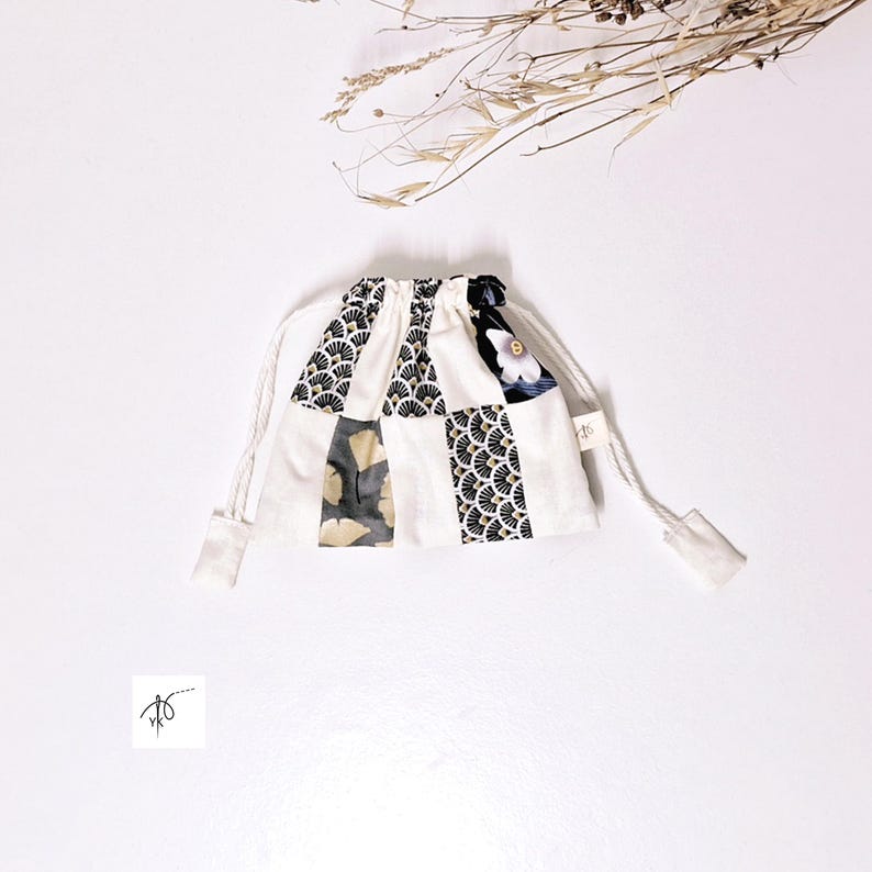 Op de afbeelding: Een kleine patchwork-zak met trekkoord. De tas is gemaakt van vierkante stoffen met patronen in zwart, goud en wit, tegen een witte achtergrond. Hij heeft een wit trekkoord en is vastgebonden met een wit koord.