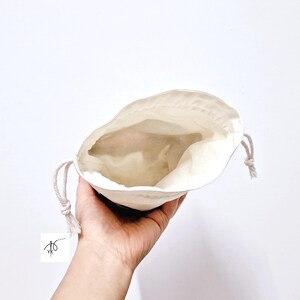 May include: An open, off-white drawstring bag held in a hand against a white background. The bag is made of a soft, natural fabric and has a simple, minimalist design. The drawstring is a light beige color.