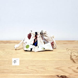 May include: A small drawstring pouch made from patchwork fabric. The fabric features a mix of floral and abstract patterns in red, white, blue, and green. The pouch is on a wooden surface.