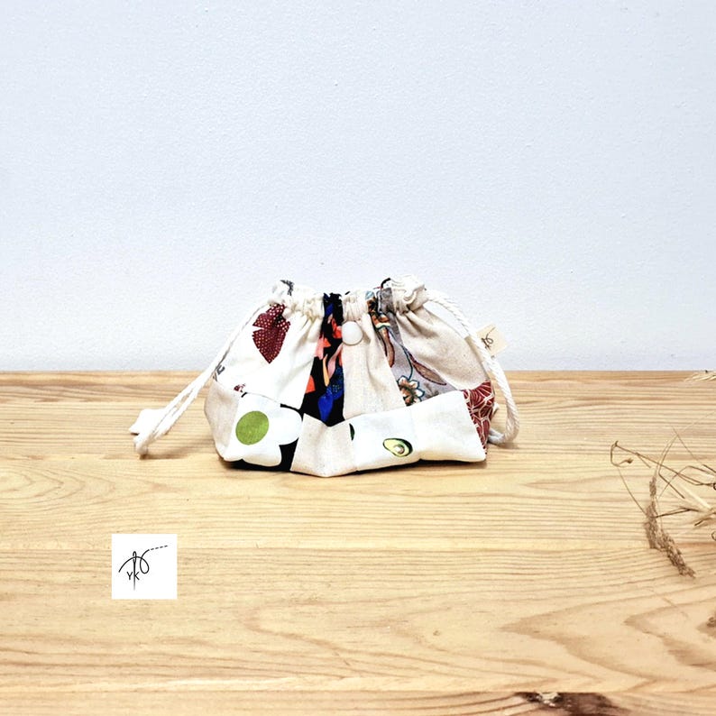 May include: A small drawstring pouch made from patchwork fabric. The fabric features a mix of floral and abstract patterns in red, white, blue, and green. The pouch is on a wooden surface.