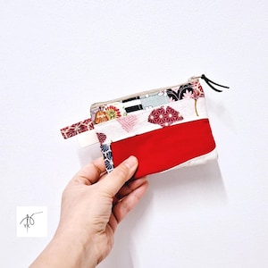 Puede incluir: Un monedero rectangular hecho a mano con un panel rojo y detalles de tela estampada. El monedero tiene cierre de cremallera y una pequeña correa de muñeca. La tela incluye diseños florales y geométricos en rojo, blanco y negro.
