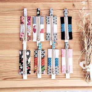 May include: A collection of handmade fabric keychains with various patterns and colors, including floral, geometric, and patchwork designs. Each keychain features a zipper closure and a metal ring for attaching keys.