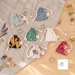 Könnte beinhalten: Eine Sammlung kleiner Stoffbeutel mit Kordelzug in verschiedenen Mustern wie Blumen, geometrischen Formen und einfarbigem Creme, begleitet von goldfarbenen Halsketten, Ringen und Trockenblumen. Ein weißes quadratisches Logo mit Nadel- und Fadensymbol ist vorhanden.