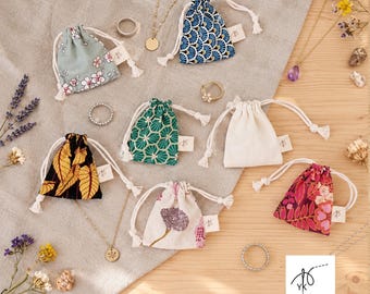 May include: A collection of small fabric drawstring pouches in various patterns including floral, geometric, and solid cream, accompanied by gold-tone necklaces, rings, and dried flowers. A white square logo with a needle and thread icon is present.