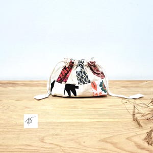 Pode incluir: Uma pequena bolsa com cordão e design de patchwork. A bolsa apresenta uma base creme com retalhos de tecido coloridos em padrões vermelhos, pretos e florais. A bolsa está sobre uma superfície de madeira.