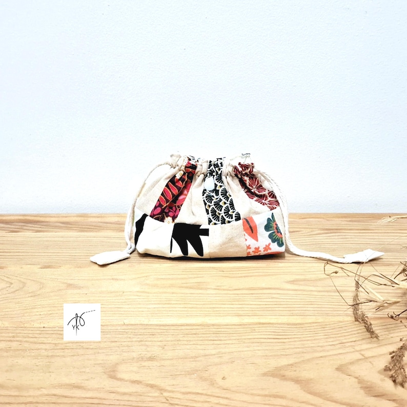 May include: A small drawstring bag with a patchwork design. The bag has a cream-coloured base with colourful fabric patches in red, black, and floral patterns. The bag is on a wooden surface.