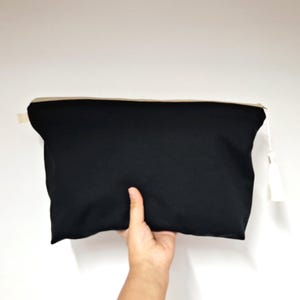 May include: A black zippered pouch with a beige zipper and a white tassel. The pouch is held by a hand against a white background. The pouch is rectangular and appears to be made of fabric.