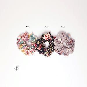 Pu&ograve; includere: Tre elastici per capelli floreali su sfondo bianco. Gli elastici sono etichettati A13, A14 e A15. A13 presenta un motivo floreale colorato, A14 ha un design floreale scuro e A15 mostra una stampa floreale tenue. Gli elastici sono in tessuto.