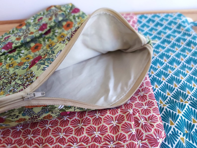 May include: Close-up of several fabric pouches with zippers. One pouch is open, revealing a cream-colored interior. The exterior fabrics feature floral and geometric patterns in shades of green, red, and teal. The zippers are beige.