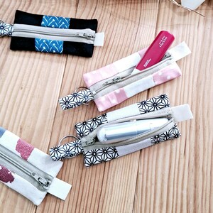 May include: Several small, rectangular fabric pouches with zippers and key rings are displayed. The pouches are in various colors, including black, pink, and white, with geometric and polka dot patterns. One pouch contains a pink lipstick tube.