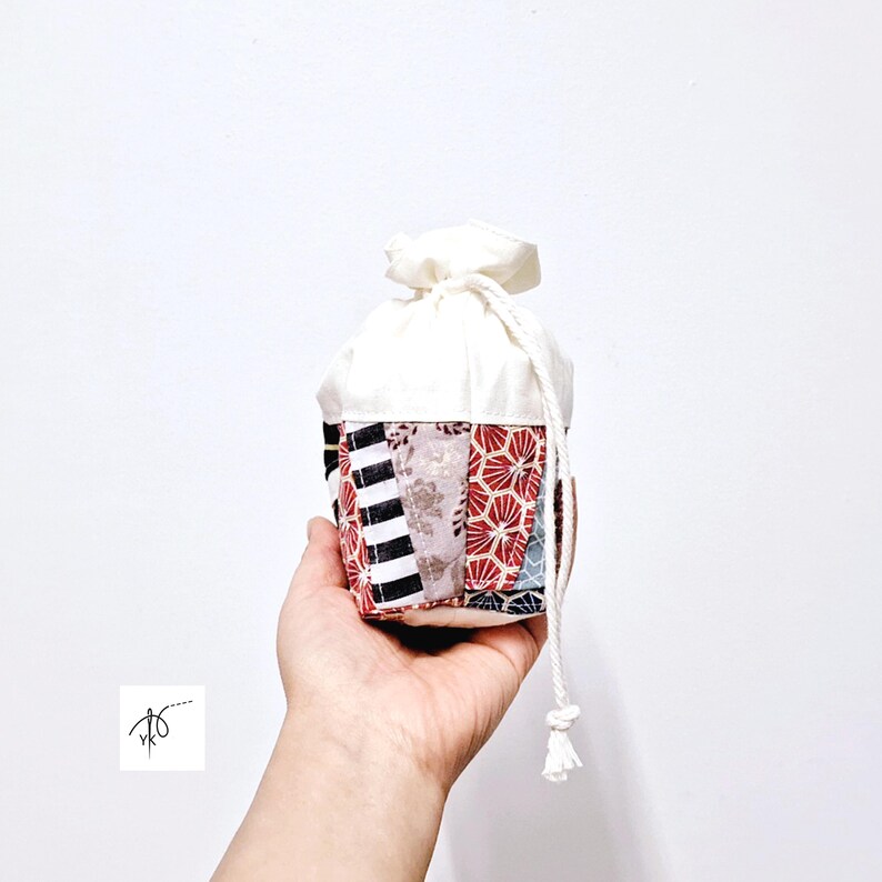 May include: A small, patchwork fabric pouch with a white drawstring closure. The pouch features a variety of patterned fabrics in red, black, and white. The top of the pouch is white, and the drawstring is made of white rope.