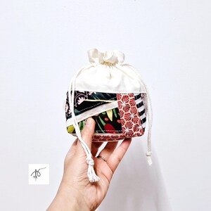 May include: A small drawstring pouch with a patchwork design. The top portion is white, while the bottom features a mix of floral, geometric, and striped fabric patterns in black, red, and green. The pouch is held by a hand.