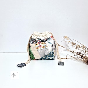 Può includere: Un sacco con coulisse patchwork con parte superiore color crema e coulisse in corda naturale. La borsa presenta una varietà di motivi in tessuto, tra cui disegni floreali, geometrici e a foglie. La base della borsa è un mix di tessuti verdi, bianchi e neri.