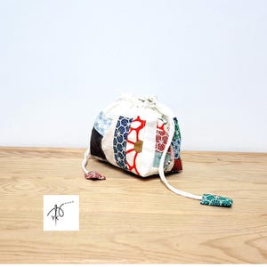 Puede incluir: Un bolso con cordón de patchwork con una parte superior blanca y paneles de tela estampada en rojo, azul y verde. Cuenta con un cierre de cordón blanco y extremos de tela decorativos. El bolso está sobre una superficie de madera.