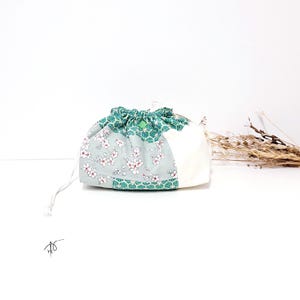 Pode incluir: Pequena bolsa com cordão e design patchwork. A bolsa apresenta secções de tecido branco, uma estampa floral com flores brancas sobre fundo azul claro e um padrão geométrico verde. Um botão verde garante o fecho com cordão.