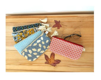May include: A collection of colorful fabric pencil cases with various patterns, including floral, geometric, and ginkgo leaf designs. Each case has a zipper closure and a tassel. A pair of eyeglasses and dried leaves are also present.
