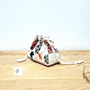 May include: A small drawstring bag with a patchwork design. The bag has a cream base with red, black, and green floral and geometric fabric patches. It is tied with a cream drawstring and rests on a wooden surface. The bag's dimensions are not visible.