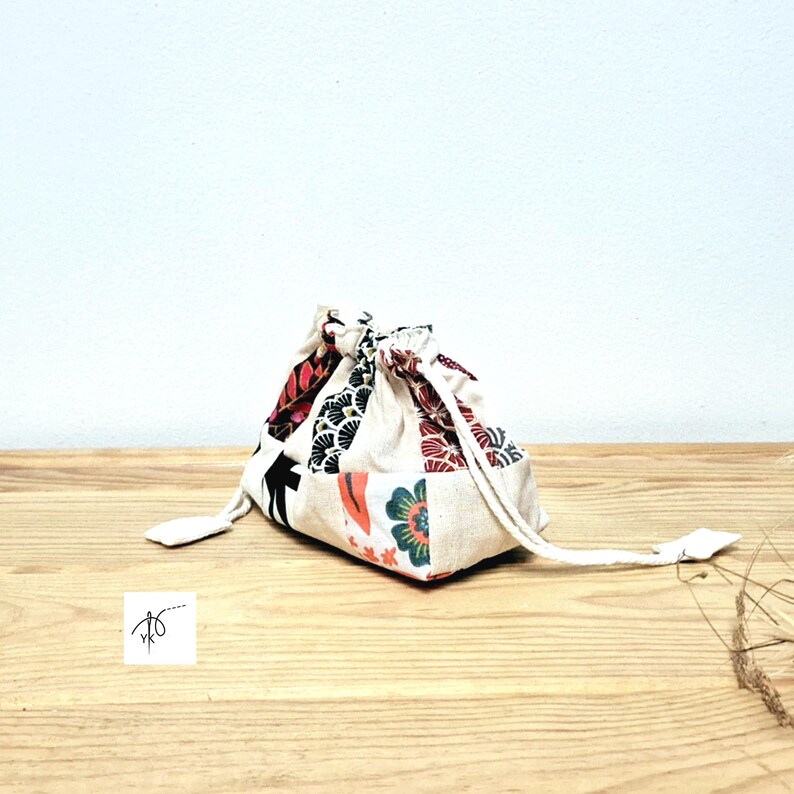 May include: A small drawstring bag with a patchwork design. The bag has a cream base with red, black, and green floral and geometric fabric patches. It is tied with a cream drawstring and rests on a wooden surface. The bag's dimensions are not visible.