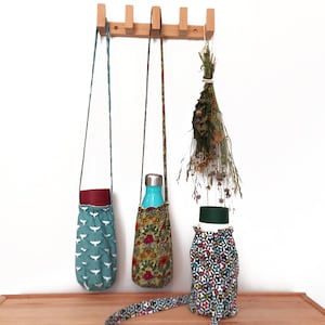 May include: Three water bottle carriers with different fabric patterns hang from a wooden rack. One is teal with white bird silhouettes, another has a floral design, and the third has a geometric pattern. A bundle of dried flowers hangs nearby.