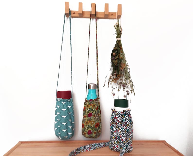 May include: Three water bottle carriers with different fabric patterns hang from a wooden rack. One is teal with white bird silhouettes, another has a floral design, and the third has a geometric pattern. A bundle of dried flowers hangs nearby.