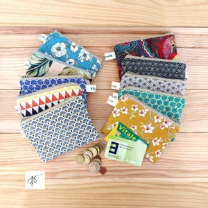 May include: A collection of ten small rectangular fabric zip pouches featuring various patterns including floral, geometric, and abstract designs. Each pouch has a small white tag labelled handmade. A few metal coins and two cards are visible.