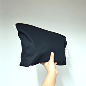 May include: A black rectangular pouch with a white zipper and a white tag. The pouch is held by a hand against a white background. The pouch appears to be made of a soft, matte fabric.