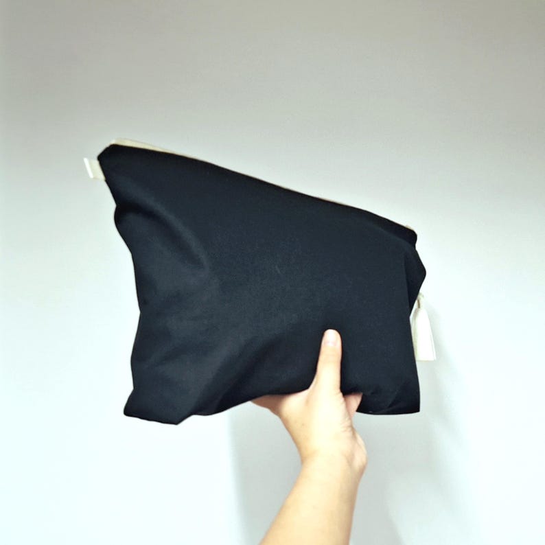 May include: A black rectangular pouch with a white zipper and a white tag. The pouch is held by a hand against a white background. The pouch appears to be made of a soft, matte fabric.