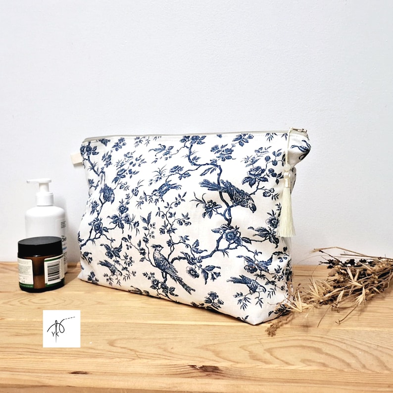 May include: A rectangular zippered pouch featuring a white fabric base with a repeating dark blue toile pattern of birds perched on flowering branches. The pouch includes a cream-colored tassel attached to the metal zipper pull on the right side.