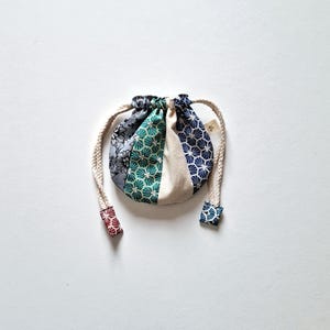 Pode incluir: Uma pequena bolsa redonda com cordão e design de patchwork. A bolsa apresenta painéis de tecido floral, geométrico e liso em tons de cinza, verde e azul. O cordão é uma corda grossa de cor creme e as extremidades são finalizadas com tecido estampado.