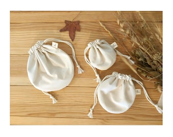 Puede incluir: Tres bolsas con cordón de color crema de diferentes tamaños están dispuestas sobre una superficie de madera. Cada bolsa tiene una parte superior fruncida y un cierre de cordón con borla. También se ven una hoja seca y algunas plantas secas.