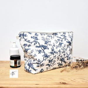 May include: A rectangular zippered pouch featuring a white fabric base with a dark blue toile pattern depicting birds perched on flowering branches. The pouch has a top zipper closure and is positioned next to a white pump bottle and a small jar.