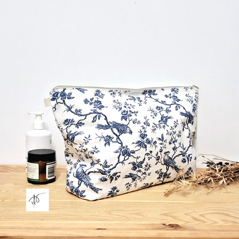 May include: A rectangular zippered pouch featuring a white fabric base with a dark blue toile pattern depicting birds perched on flowering branches. The pouch has a top zipper closure and is positioned next to a white pump bottle and a small jar.