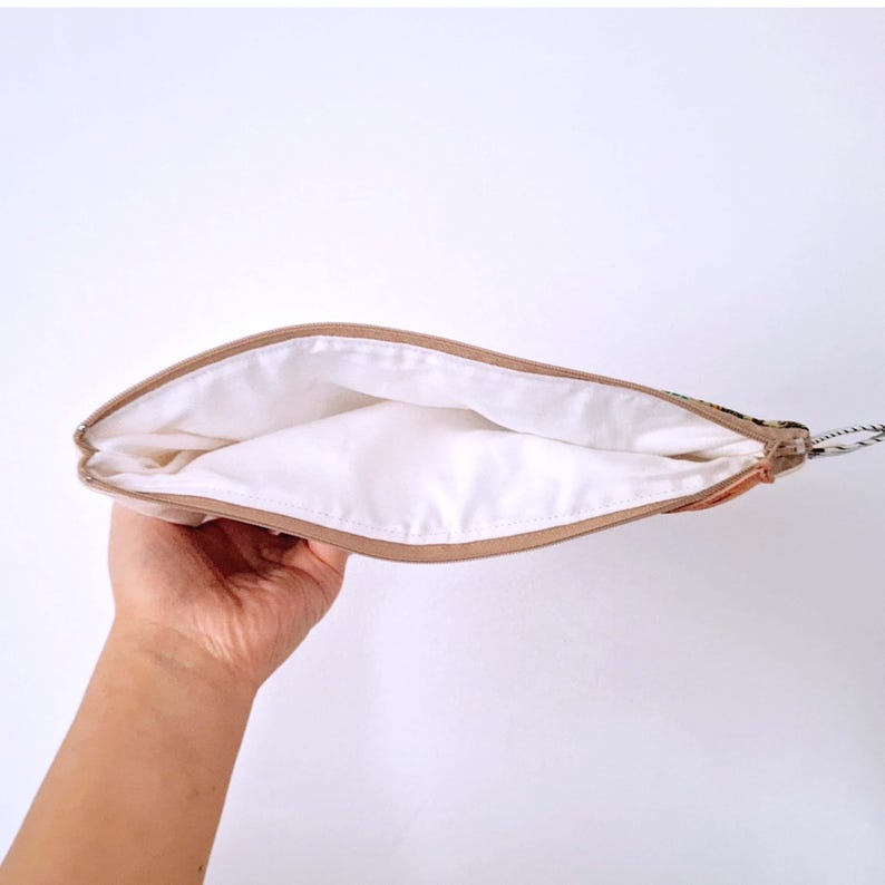 Op de afbeelding: Een beige etui met ritssluiting en een witte binnenkant wordt opengehouden, waardoor de binnenkant zichtbaar is. De etui heeft een bruine rand en een kleine lus met een touwtje eraan. De achtergrond is effen wit.