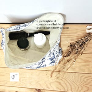 May include: A white zippered pouch with a blue floral pattern containing a black hairbrush, a black jar, and a white bottle. The interior text reads: Big enough to fit cosmetics and hair brush and still have plenty of room.